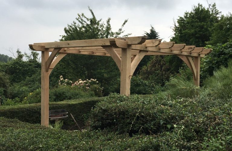 A wooden pergola with curved braces over a hedge-lined garden, featuring flowering bushes and trees beyond under a cloudy sky.