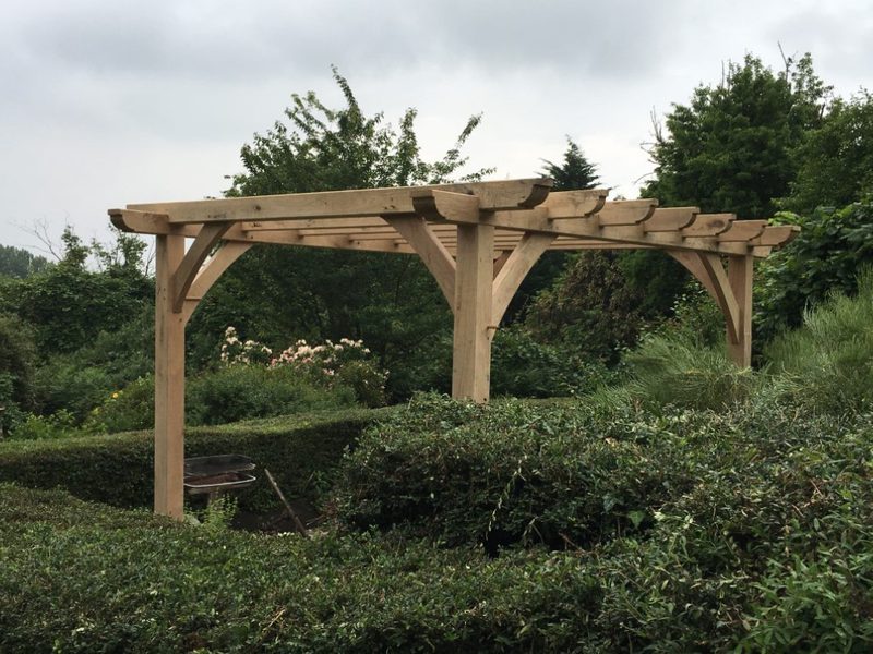 A wooden pergola with curved braces over a hedge-lined garden, featuring flowering bushes and trees beyond under a cloudy sky.