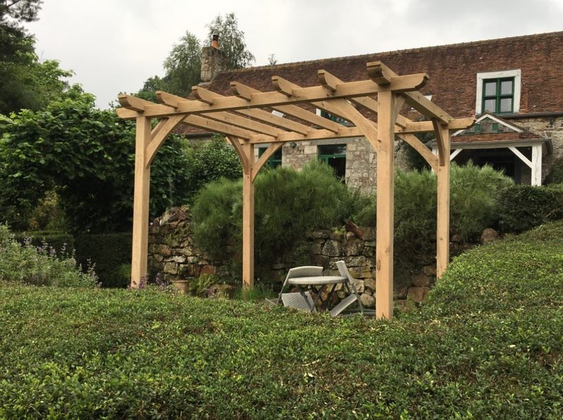 Wooden garden pergola with a folding chair and small table beneath, in a lush yard with hedges and a stone house in the background.