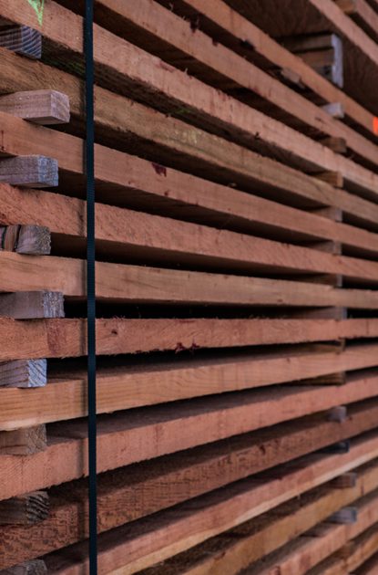 Rows of brown wooden planks stacked and strapped in a lumber yard, with visible grain and orange tags along the edges, receding into the distance.